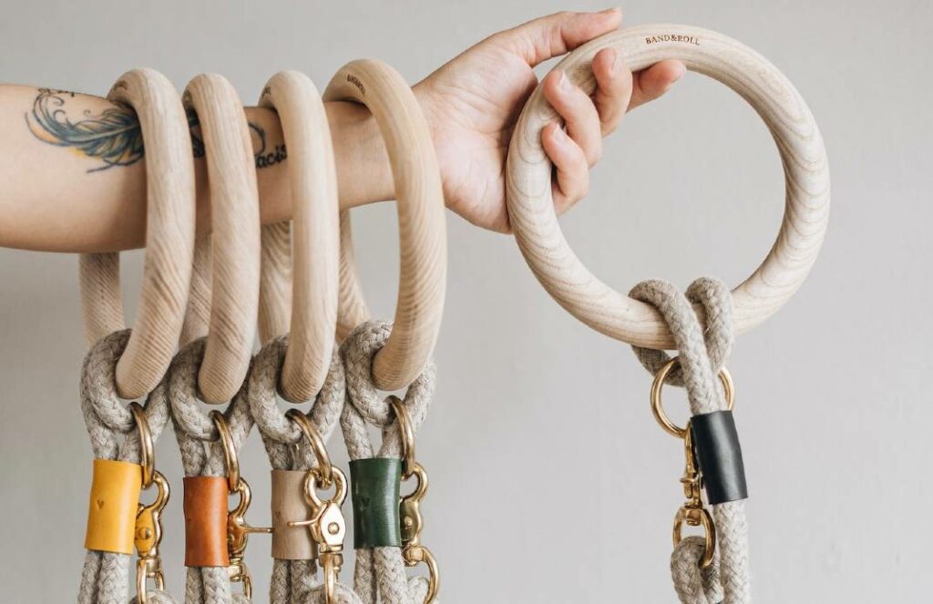 Tattooed arm holding multiple hemp dog leashes with polished dead ash wood handles and vegetable-tanned leather fittings in different colors.