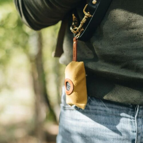 Close-up of mustard-colored waste bag dispenser by Band & Roll attached to leash O-ring, worn by man outdoors.